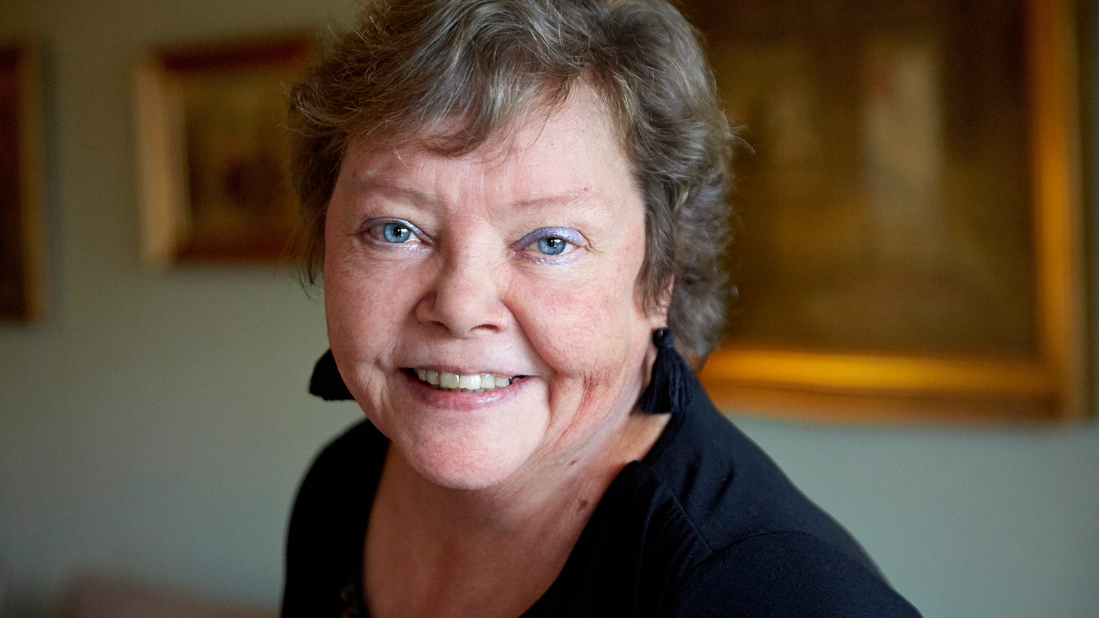 Smilende ældre kvinde med kort gråt hår ser direkte ind i kameraet i et varmt oplyst indendørsmiljø.