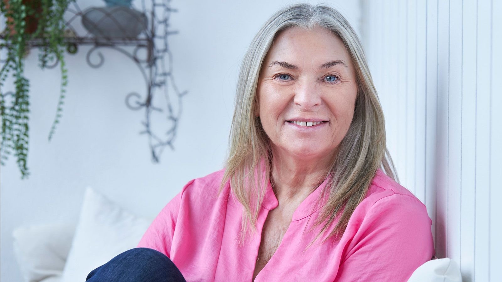 Smilende kvinde med gråt hår og lyserød bluse sidder afslappet i et lyst hjem.